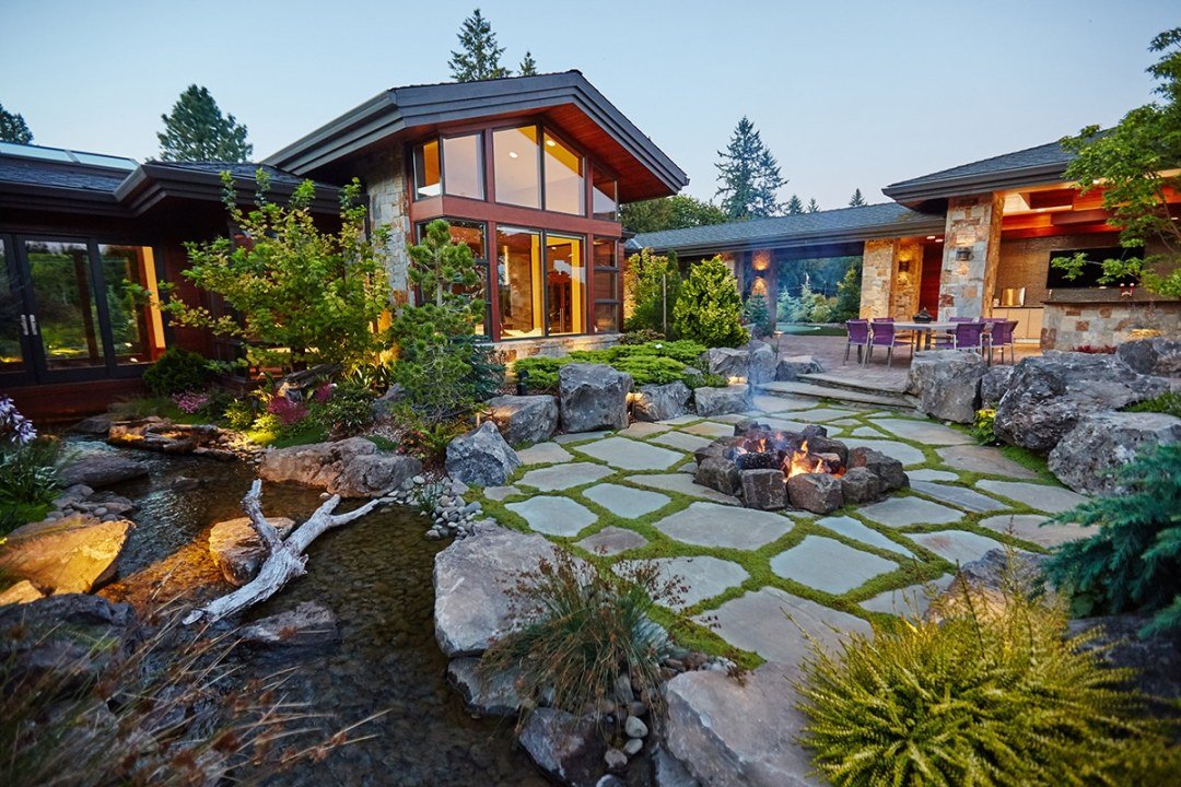 PNW Dream Home at Dusk - 7 Dees Landscaping Dusk landscape with paver patio, firepit, and stream with boulders in front of a large house - 7 Dees Landscaping